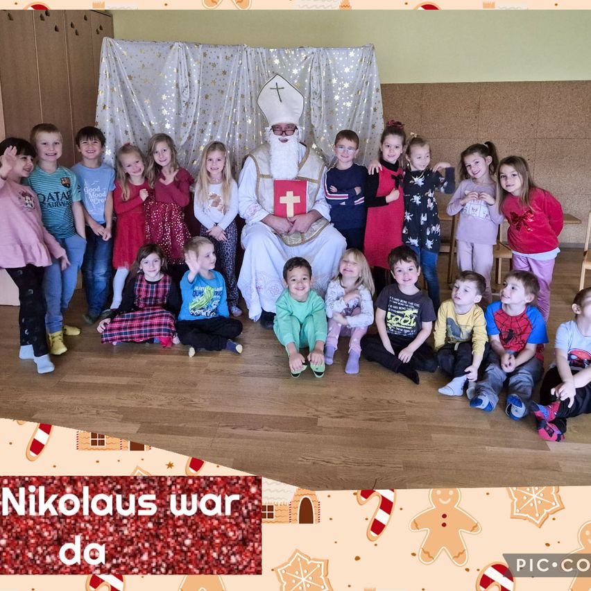 Eine Gruppe von Kindern ist um einen Mann versammelt, der als Sankt Nikolaus gekleidet ist und ein rotes Buch mit einem Kreuz in der Hand hält. Sie lächeln und posieren für ein Foto, wobei einige auf dem Boden sitzen und andere stehen. Der Raum ist mit Ornamenten geschmückt und der Text lautet Nikolaus war da.