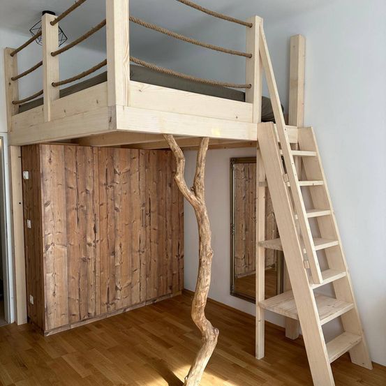 Ein hölzerne Hochbett mit einer Leiter und einem Seilgeländer, neben einer Holzwand und einem Spiegel. Der Raum verfügt über Holzboden und natürliches Licht.
