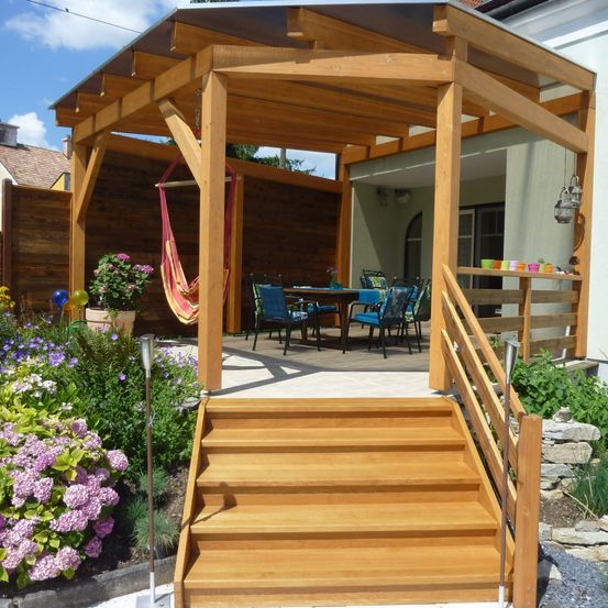 Eine Holzterrasse mit einem Pavillon, einem Tisch und Stühlen. Ein Hängesessel hängt von der Decke. Ein Garten mit bunten Blumen und einer Lampe befindet sich links.