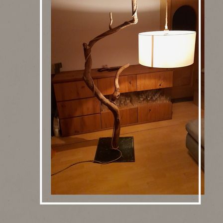 Ein einzigartiger Stehlampe aus Holzzweigen steht in einem Zimmer mit einem Lampenschirm, der sanftes Licht spendet. Dahinter befindet sich eine Holzkommode und eine Couch.