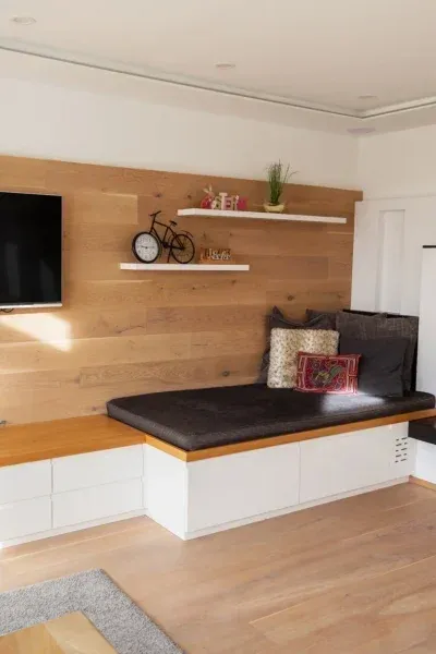 Ein modernes Wohnzimmer mit Eckbett, Holzwand, Fernseher und Regalen mit Dekorationsartikeln. Der Boden ist aus Holz und der Raum gut beleuchtet.