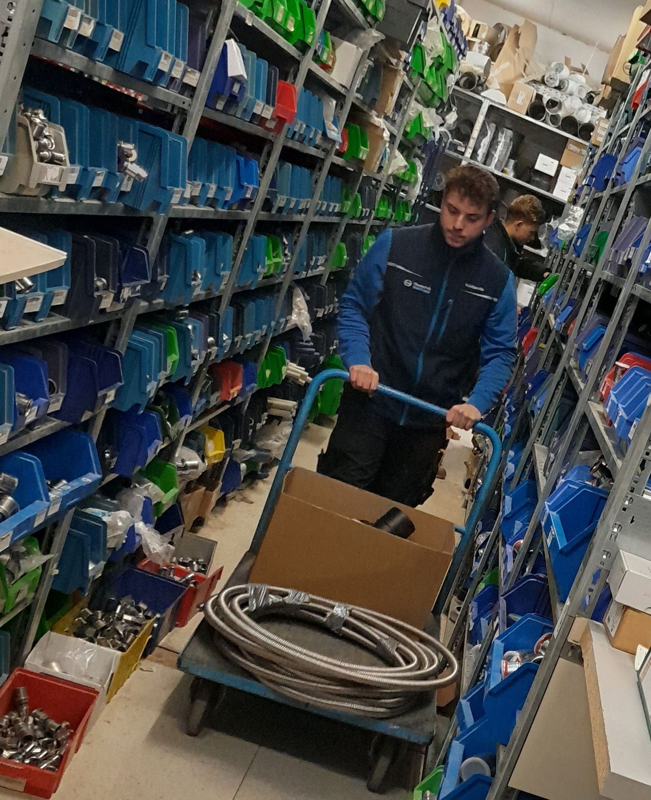 Ein Mann in einer blauen Jacke schiebt einen Wagen mit einem Schlauch durch ein Lager, das mit blauen Lagerboxen gefüllt ist.