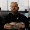 Ein bärtiger Mann mit schwarzem Shirt und sichtbaren Tattoos an den Armen steht mit verschränkten Armen.
