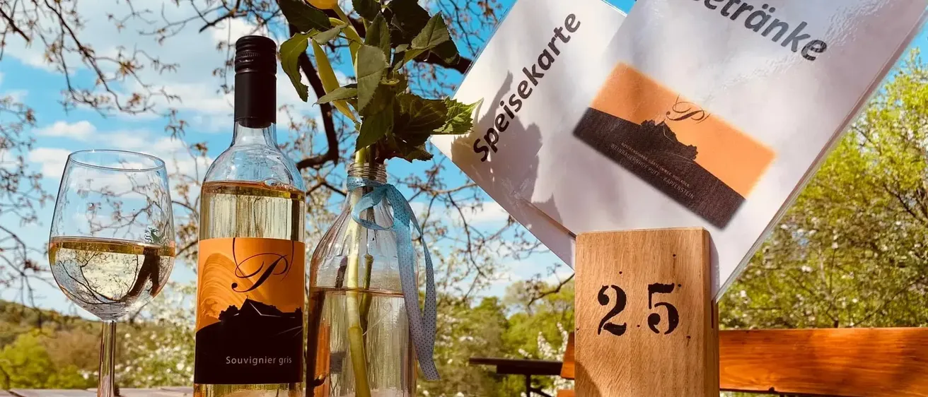 Eine Flasche Weißwein, ein Weinglas, ein Blumenvase und eine Tischnummer 25 mit zwei Broschüren mit den Titeln 'Speisekarte' und 'Getränke' stehen auf einem Holztisch.
