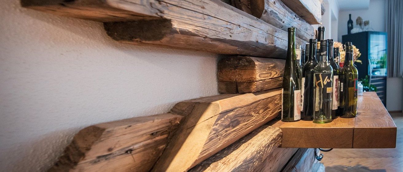 Ein Holzregal mit mehreren Weinflaschen darauf. Die Wand dahinter besteht aus großen Holzbalken. Eine Lampe ist an der Decke angebracht.