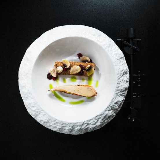 Eine weiße Schüssel mit strukturierter Oberfläche enthält ein Gericht mit einem Fruchtstück, Pilzen und roten Beeren, mit grüner Sauce.