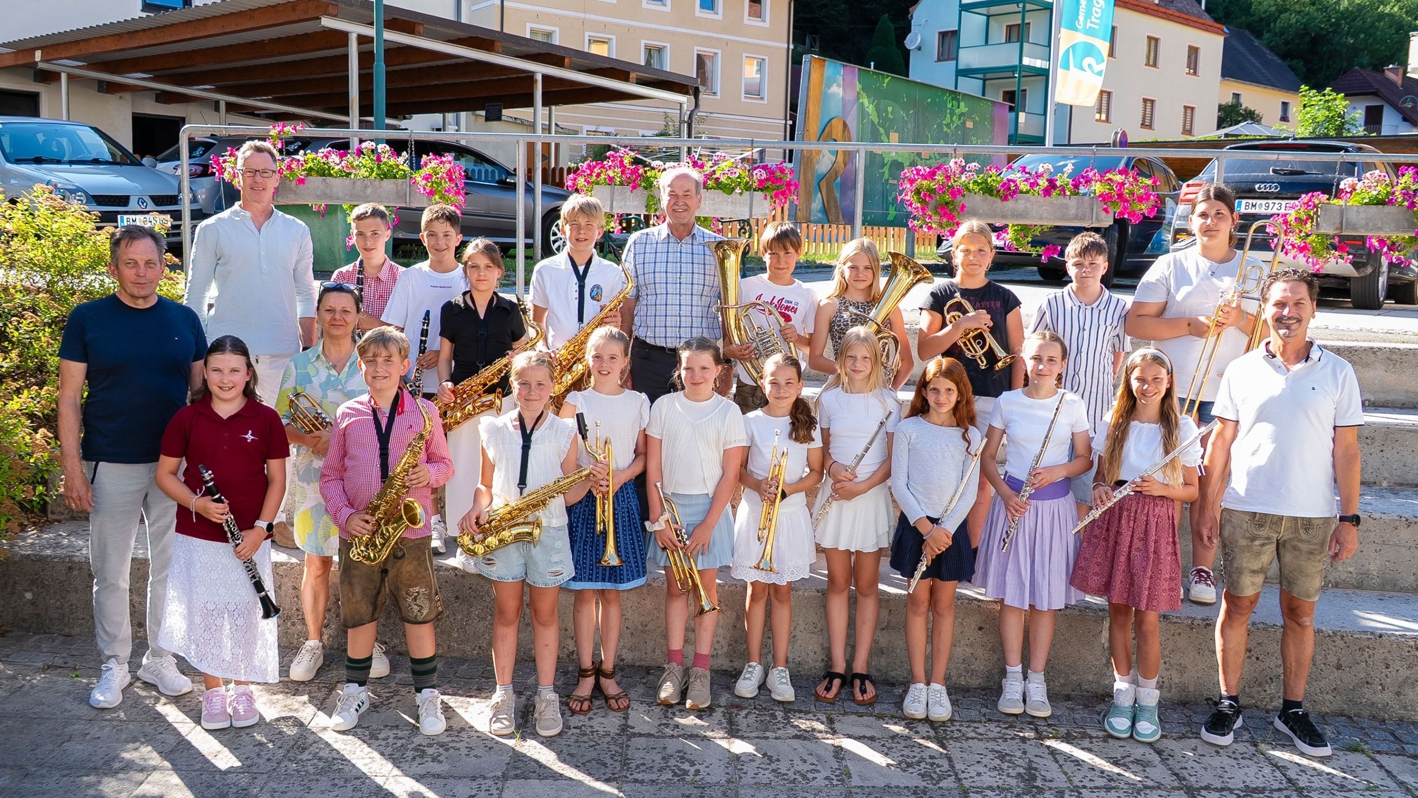 Eine Gruppe von Kindern und Erwachsenen posiert mit verschiedenen Musikinstrumenten, darunter Saxophone, Trompeten und Tuben, vor einem Gebäude mit einem Wandbild.