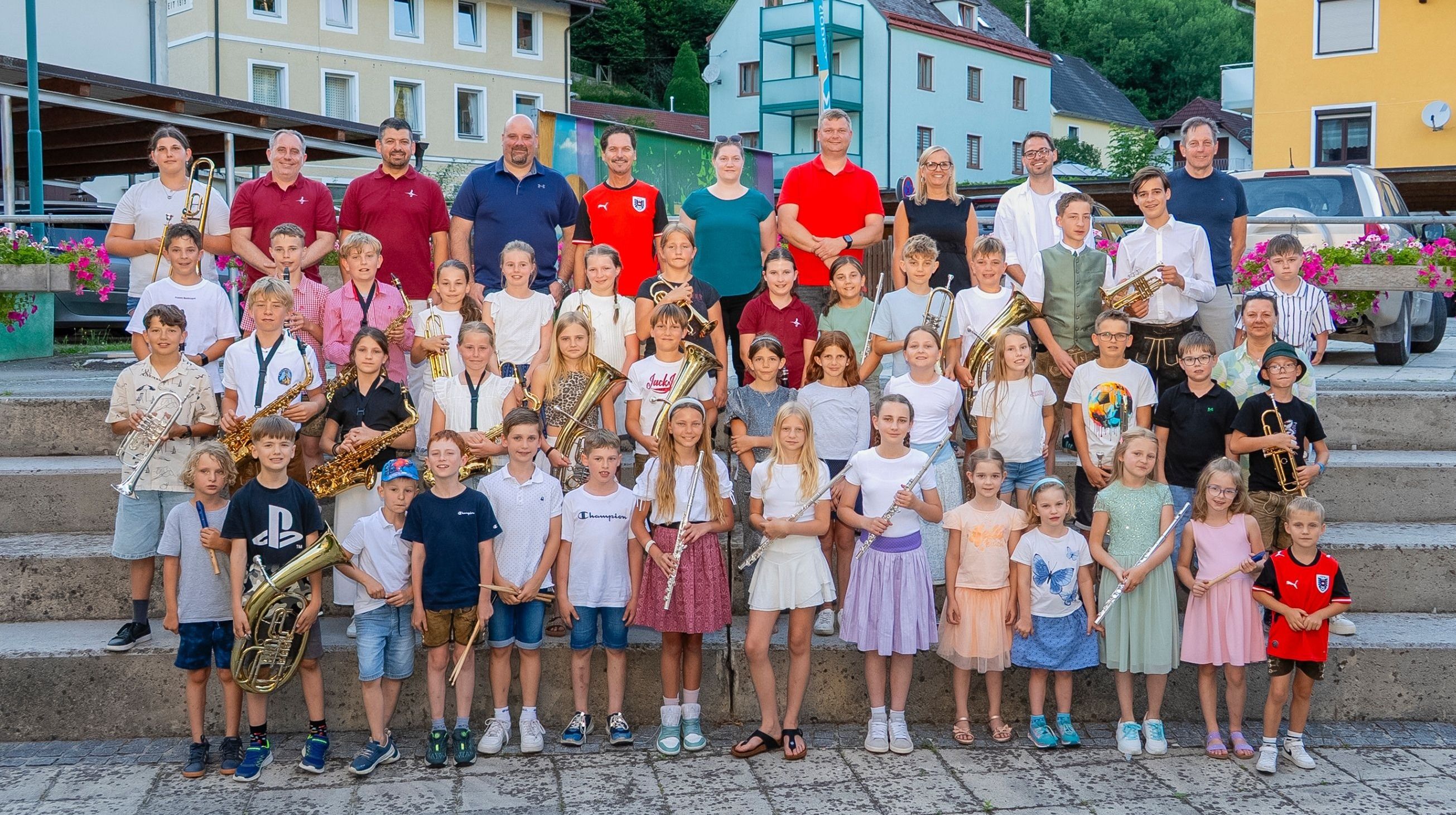 Eine Gruppe junger Musiker, Jungen und Mädchen, steht zusammen und hält verschiedene Instrumente, wobei Erwachsene hinter ihnen stehen. Sie stehen vor einem Gebäude mit einem farbenfrohen Wandbild.
