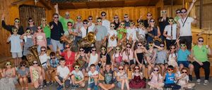 Eine Gruppe von Musikern, sowohl Erwachsene als auch Kinder, posiert für ein Foto, während sie Sonnenbrillen tragen und verschiedene Musikinstrumente halten. Sie befinden sich in einem Raum mit Holzwänden und einer grünen Decke.