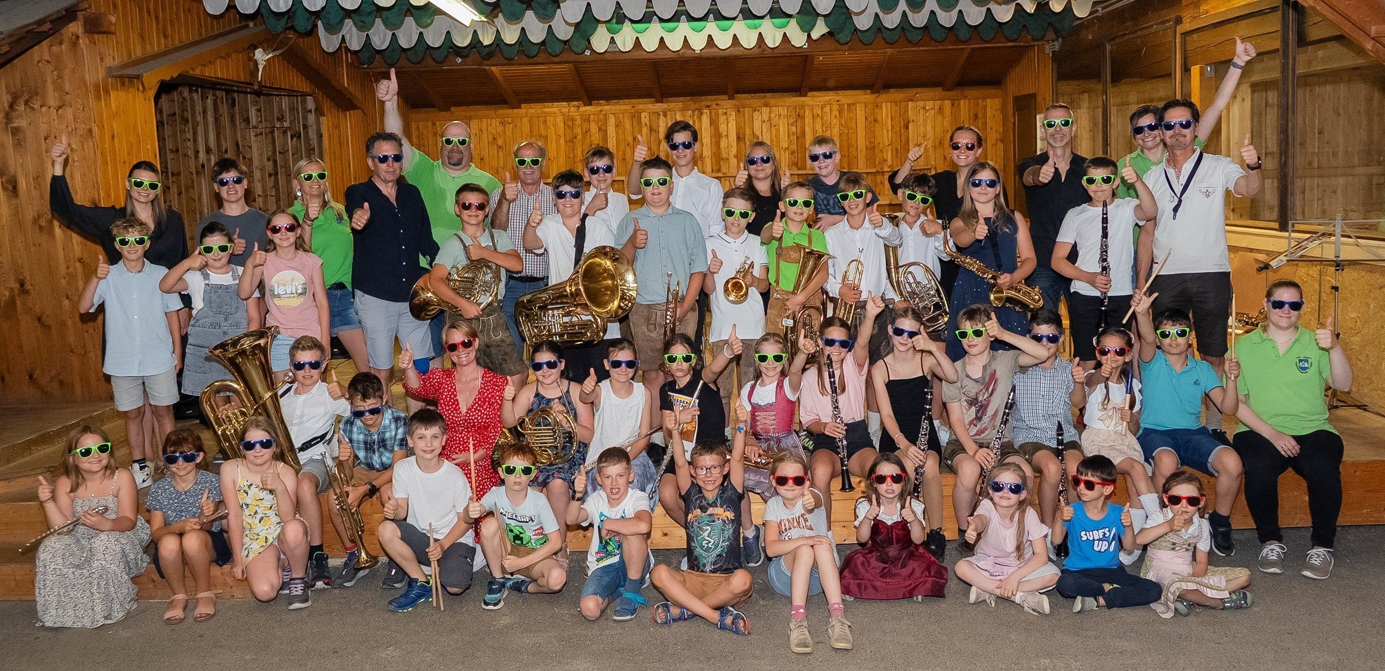 Eine Gruppe von Musikern, sowohl Erwachsene als auch Kinder, posieren für ein Foto und tragen Sonnenbrillen und halten Musikinstrumente wie Trompeten, Posaunen und Flöten. Sie alle lächeln und geben Daumen hoch.