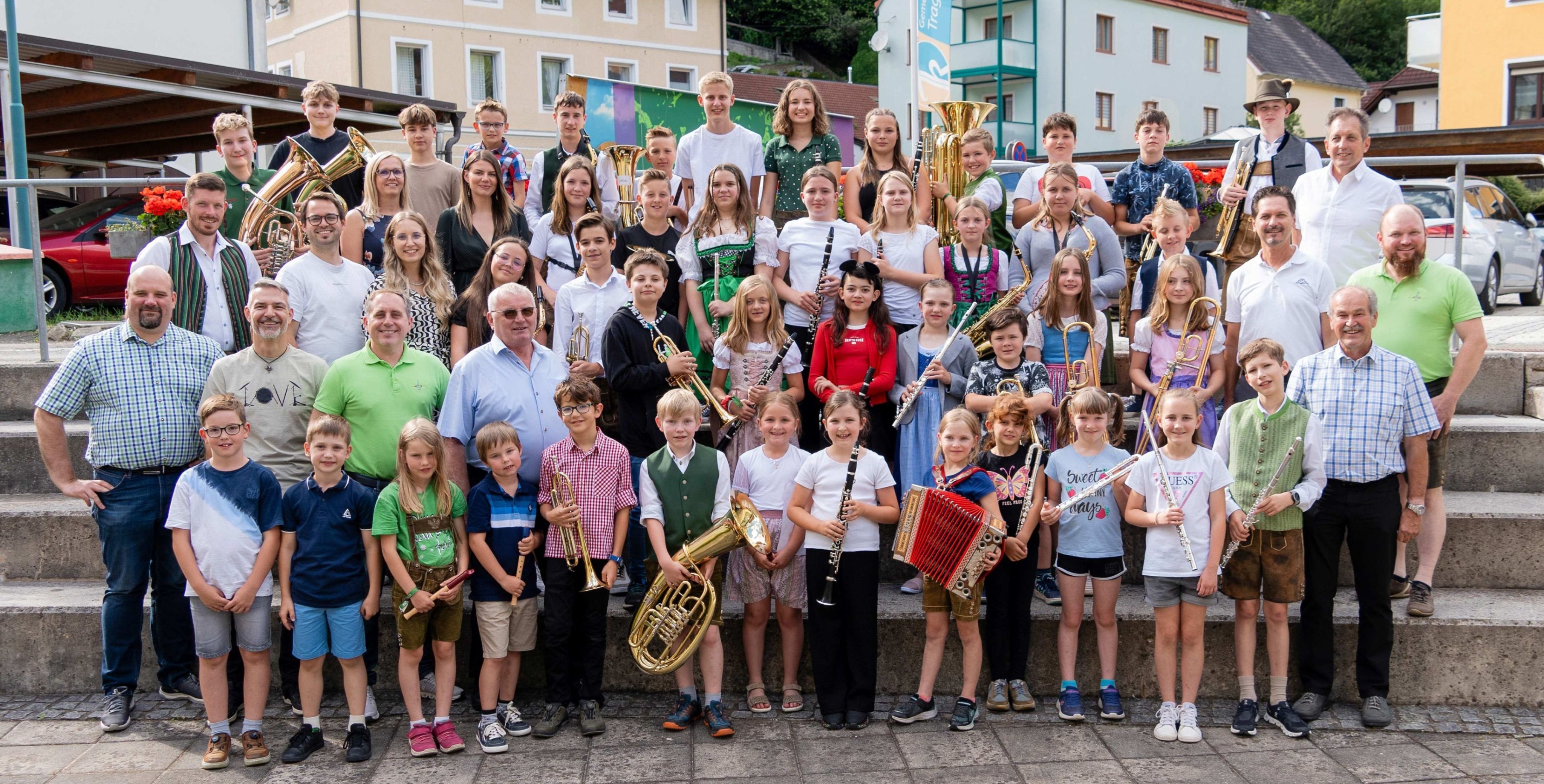 Eine Gruppe junger Musiker posiert mit ihren Instrumenten vor einem Gebäude. Einige tragen traditionelle Kleidung, andere tragen Freizeitkleidung.