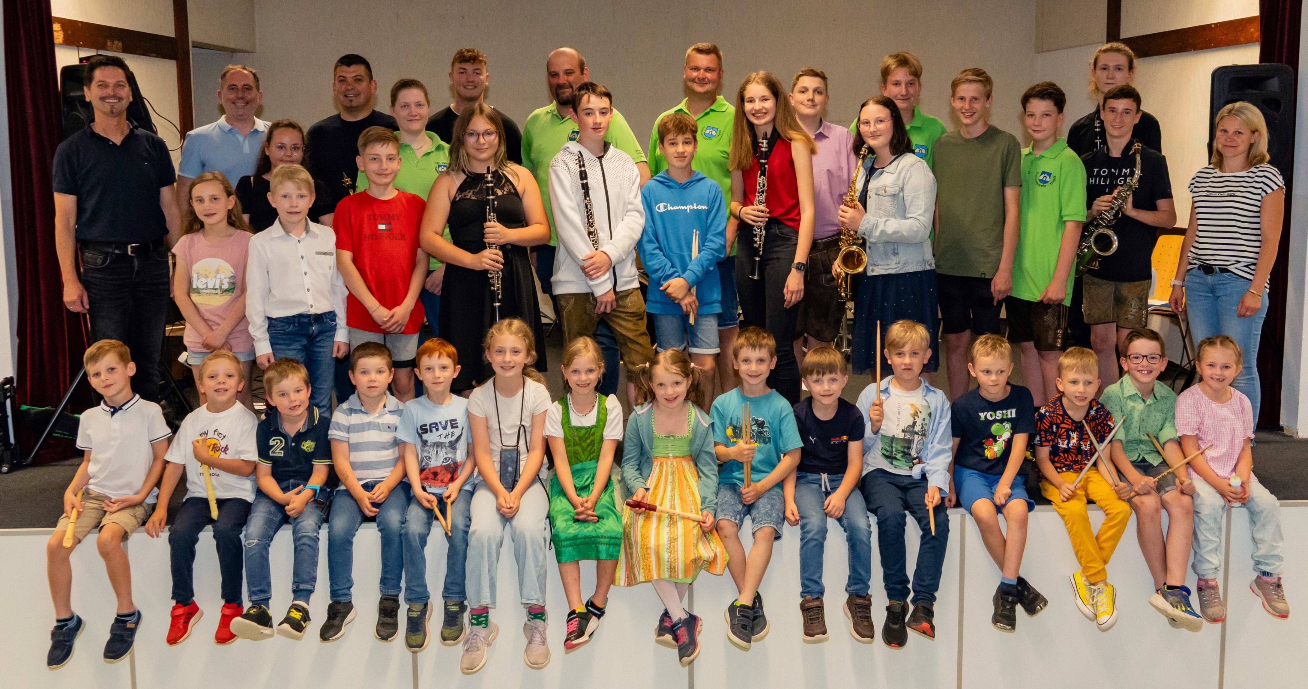 Ein Gruppenfoto einer Musikband mit Kindern und Erwachsenen. Einige halten Musikinstrumente. Die Kinder sind in Freizeitkleidung gekleidet.
