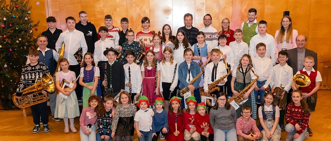 Eine Gruppenaufnahme von Kindern und Erwachsenen, alle mit Musikinstrumenten und festlicher Kleidung. Sie stehen und sitzen in einem Halbkreis auf einer Bühne.