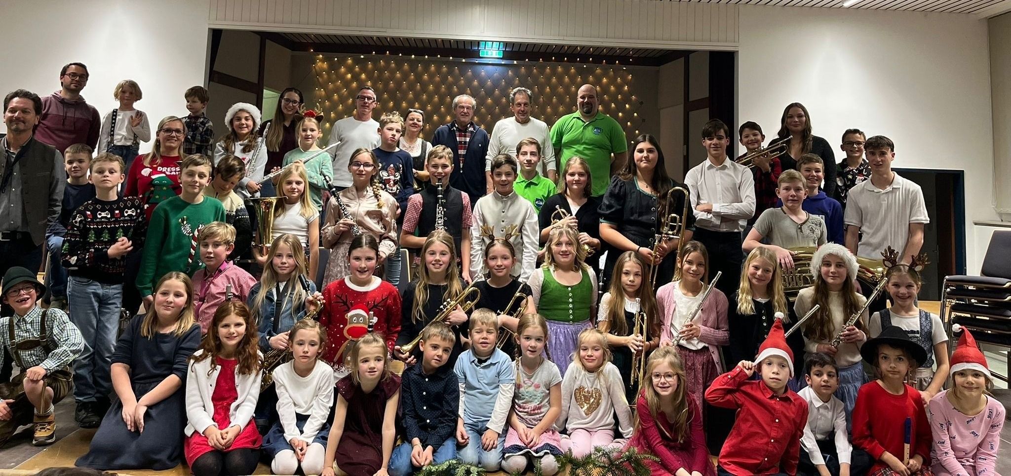 Eine Gruppe von Kindern und Erwachsenen posiert für ein Foto, alle halten Musikinstrumente wie Trompeten, Saxophone und Posaunen. Sie sind drinnen, mit einer mit Lichtern geschmückten Wand im Hintergrund.