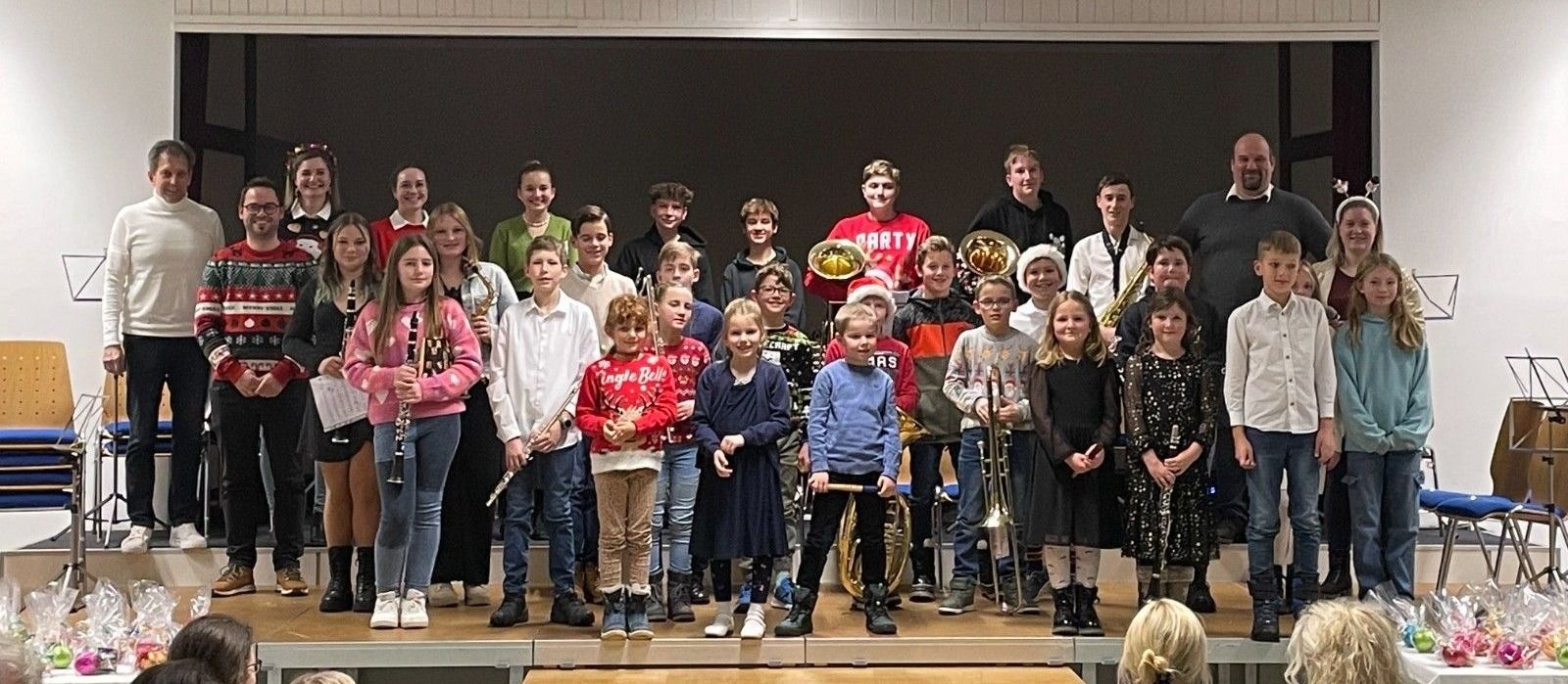 Eine Gruppe junger Musiker steht auf einer Bühne und hält verschiedene Instrumente. Einige tragen Mützen und Turnschuhe. Die Bühne hat einen Holzboden und einen schwarzen Hintergrund.