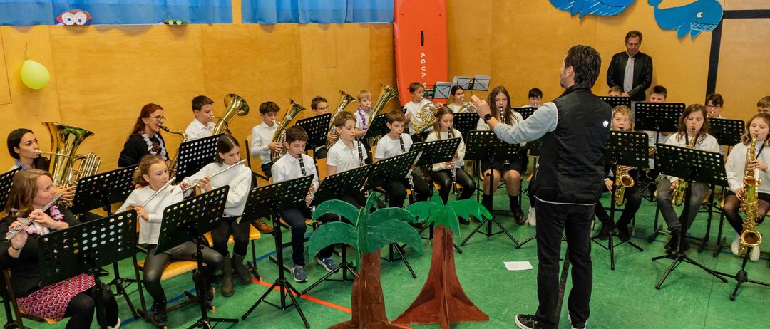 Eine Band von Kindern spielt verschiedene Instrumente in einem Klassenzimmer, das mit Meerestieren und einem Surfbrett dekoriert ist. Ein Dirigent steht davor und leitet die Aufführung.