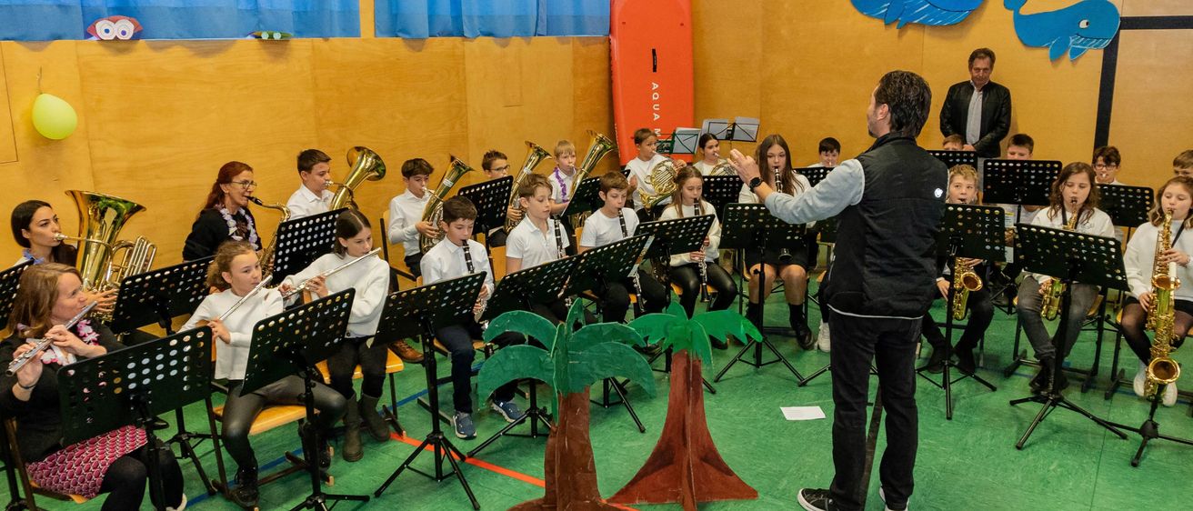Eine Band von Kindern spielt verschiedene Instrumente in einem Klassenzimmer, das mit Meerestieren und einem Surfbrett dekoriert ist. Ein Dirigent steht davor und leitet die Aufführung.