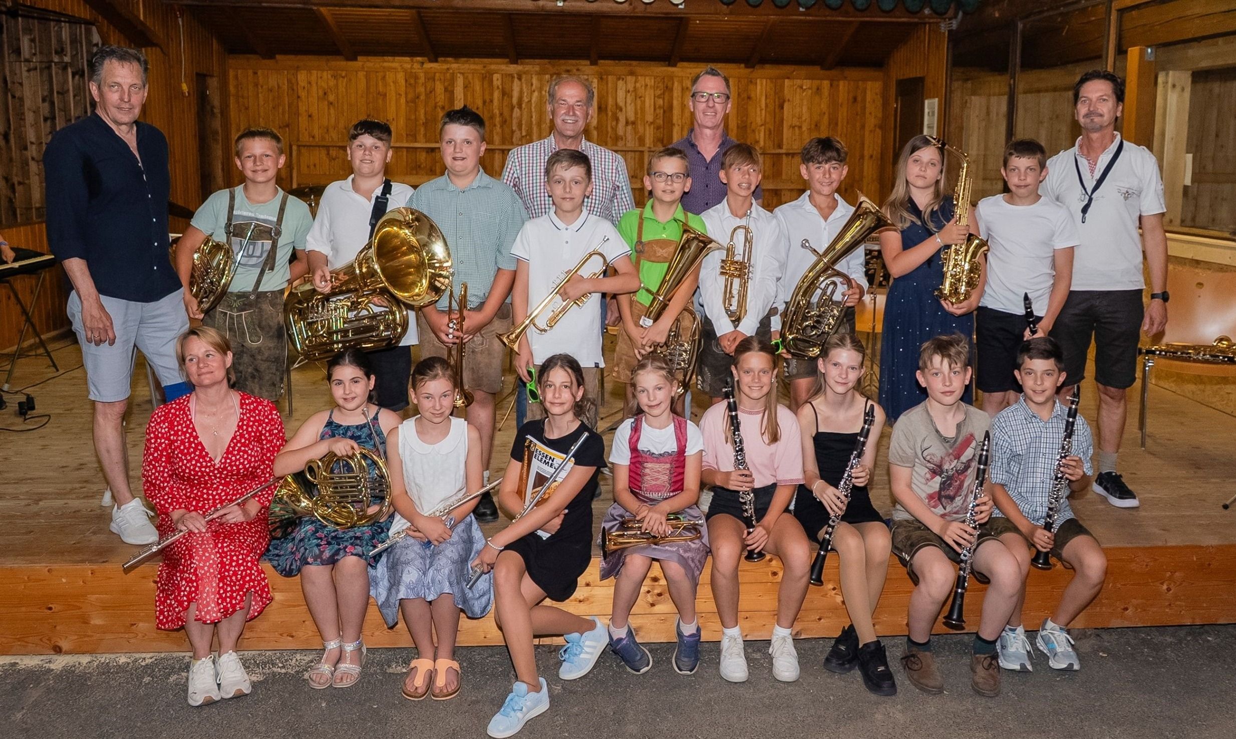 Eine Gruppe junger Musiker in einem Musikzimmer, die für ein Foto mit verschiedenen Instrumenten wie Trompeten, Posaunen und Klarinetten unter einer Holzdecke posiert.