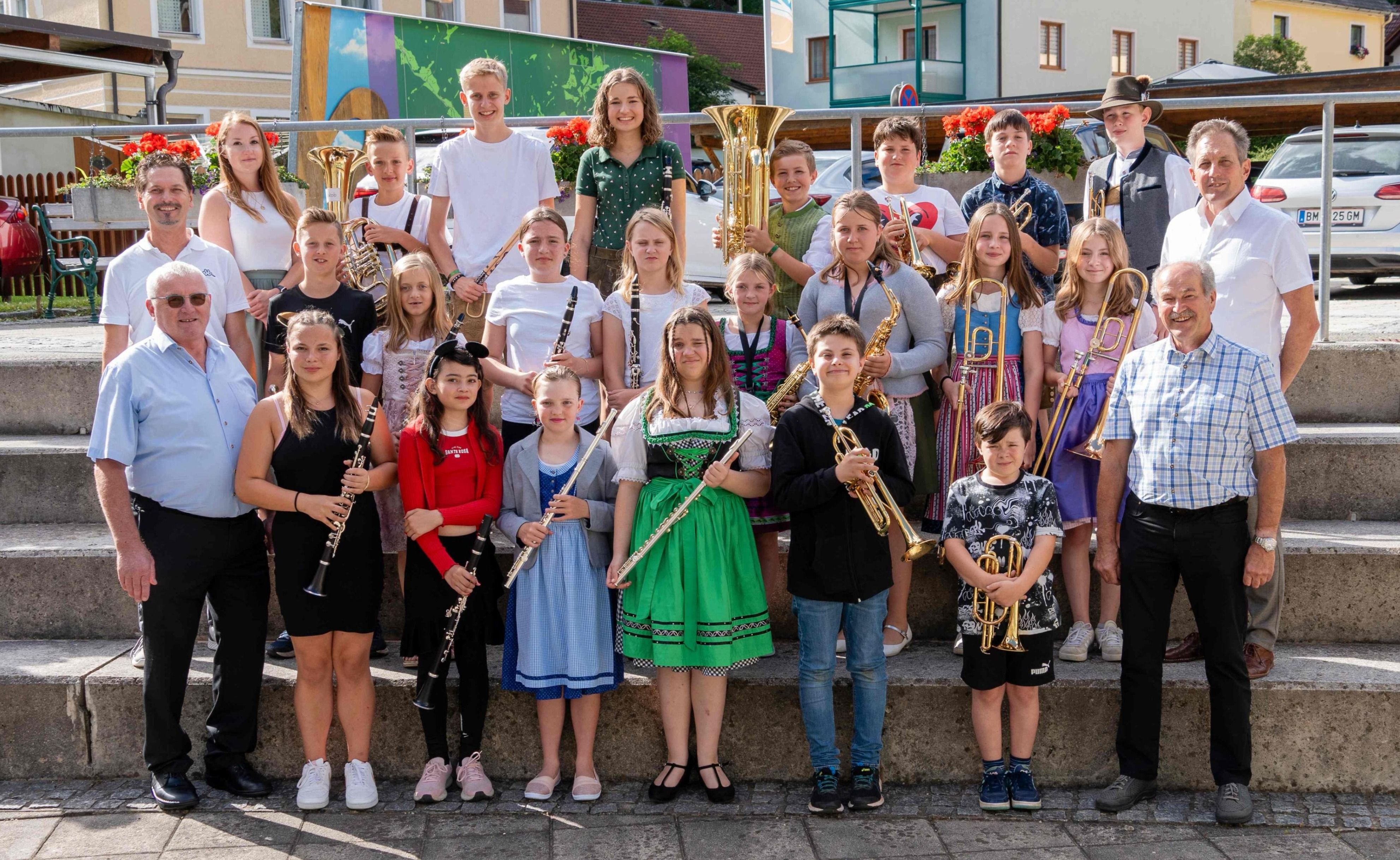 Eine Gruppe von Kindern in traditioneller und lässiger Kleidung, die auf einer Betonplattform mit Musikinstrumenten stehen, einige halten Trompeten, Flöten und Saxophone.