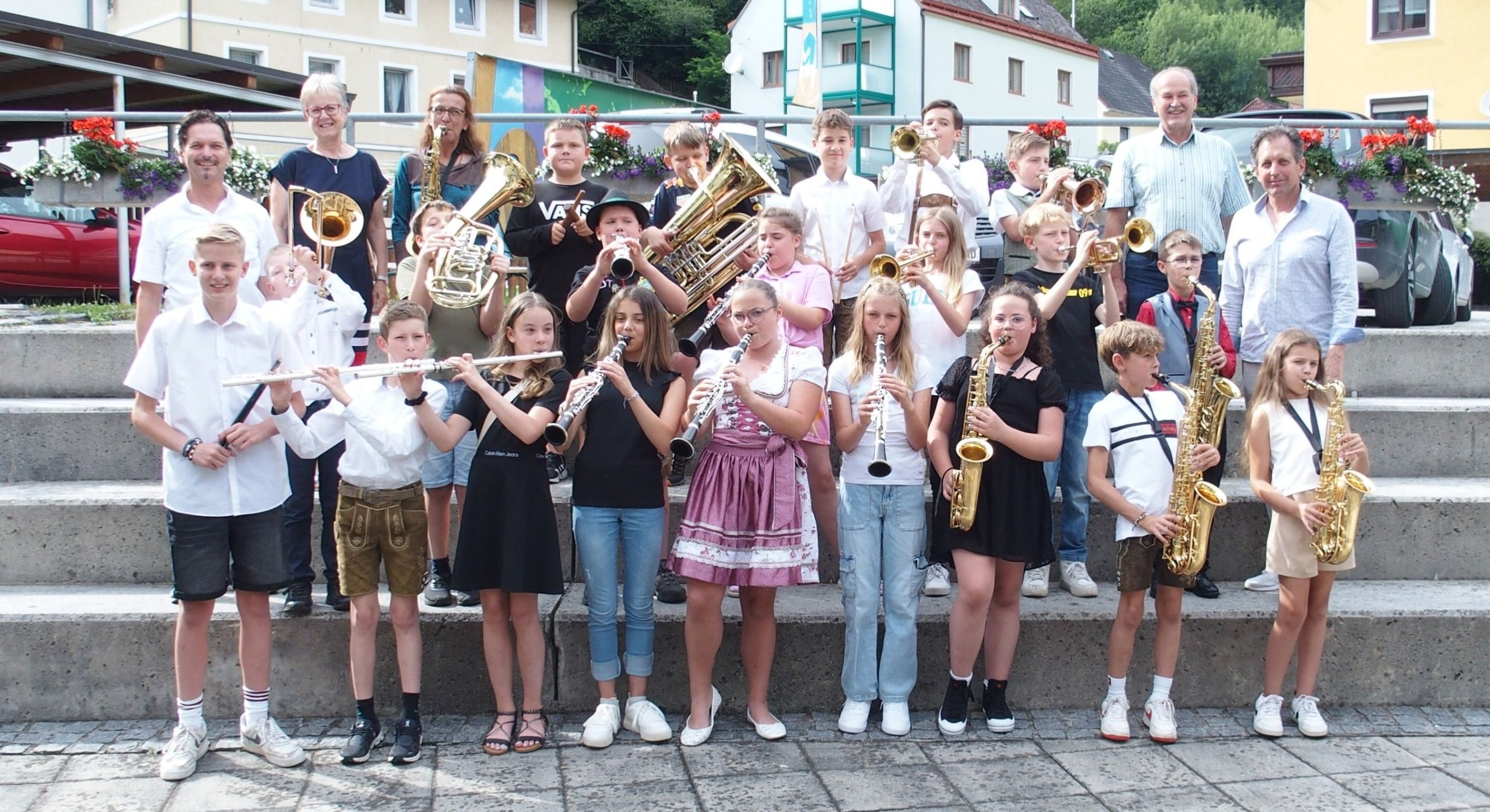 Eine Gruppe von Kindern posiert für ein Foto, während sie im Freien Musikinstrumente spielen. Einige halten Trompeten und andere Saxophon. Sie stehen auf Betonstufen.