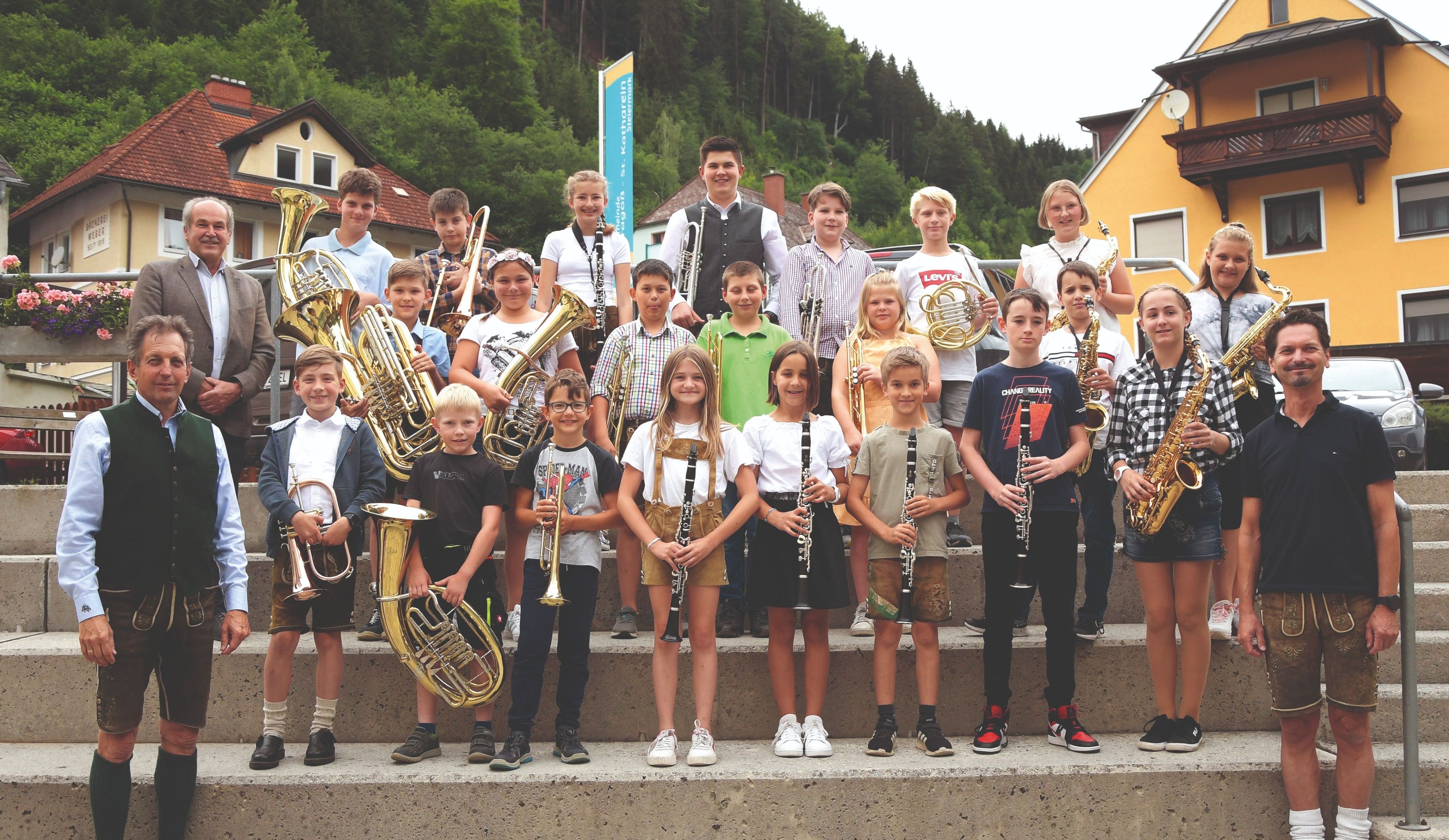 Eine Gruppe von Kindern und Jugendlichen steht mit verschiedenen Musikinstrumenten wie Trompeten, Saxophonen und Klarinetten auf einer Treppe. Sie sind in lässiger Kleidung gekleidet, einige tragen traditionelle Gewänder. Sie lächeln für das Foto mit einer malerischen Kulisse von Häusern und Bäumen.
