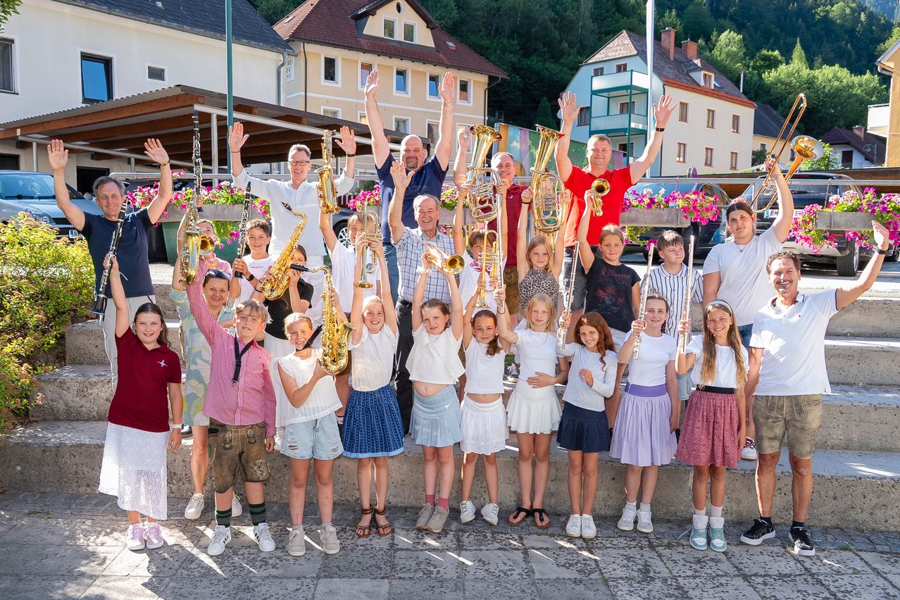 A group of children and adults are posing for a photo while holding musical instruments in a town square.