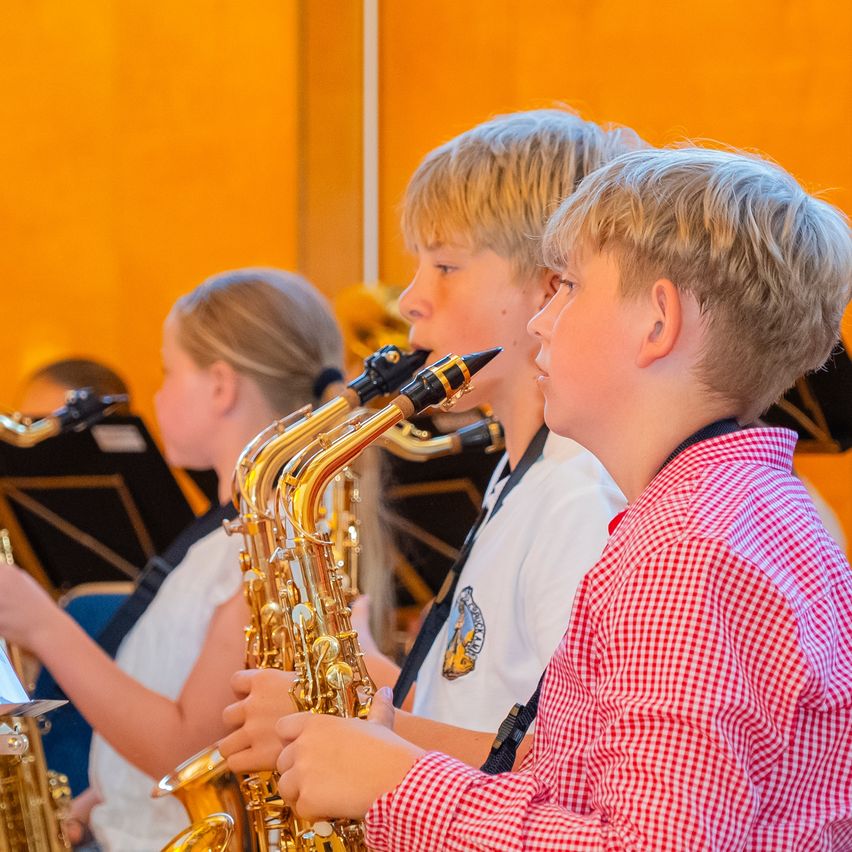 Three children in a school band are playing saxophones. The boy in the foreground wears a red checkered shirt.