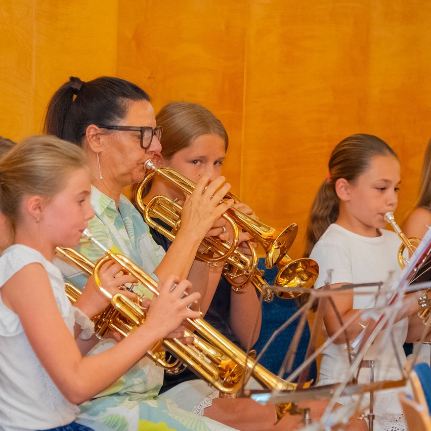 A group of young girls and an adult woman are playing trumpets in a room. The adult woman is wearing glasses.