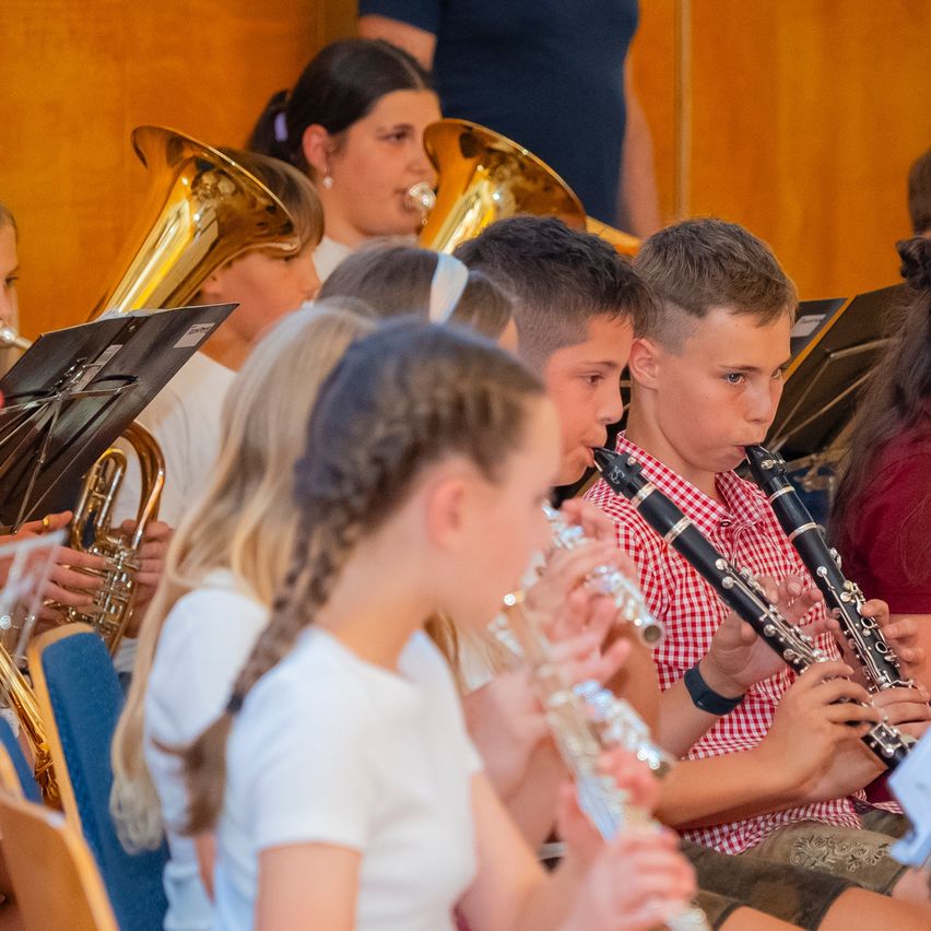 A group of young musicians, including boys and girls, are playing various instruments such as trumpets, flutes, and clarinets in a well-lit room.