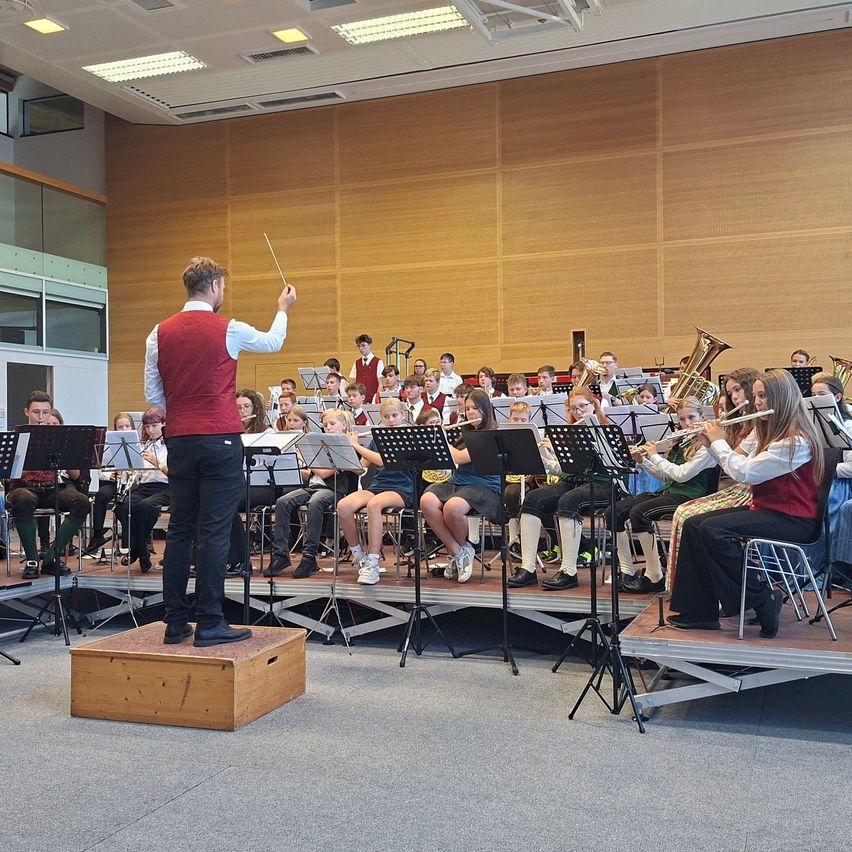A conductor leads a large orchestra in a well-lit room, with musicians playing various instruments.