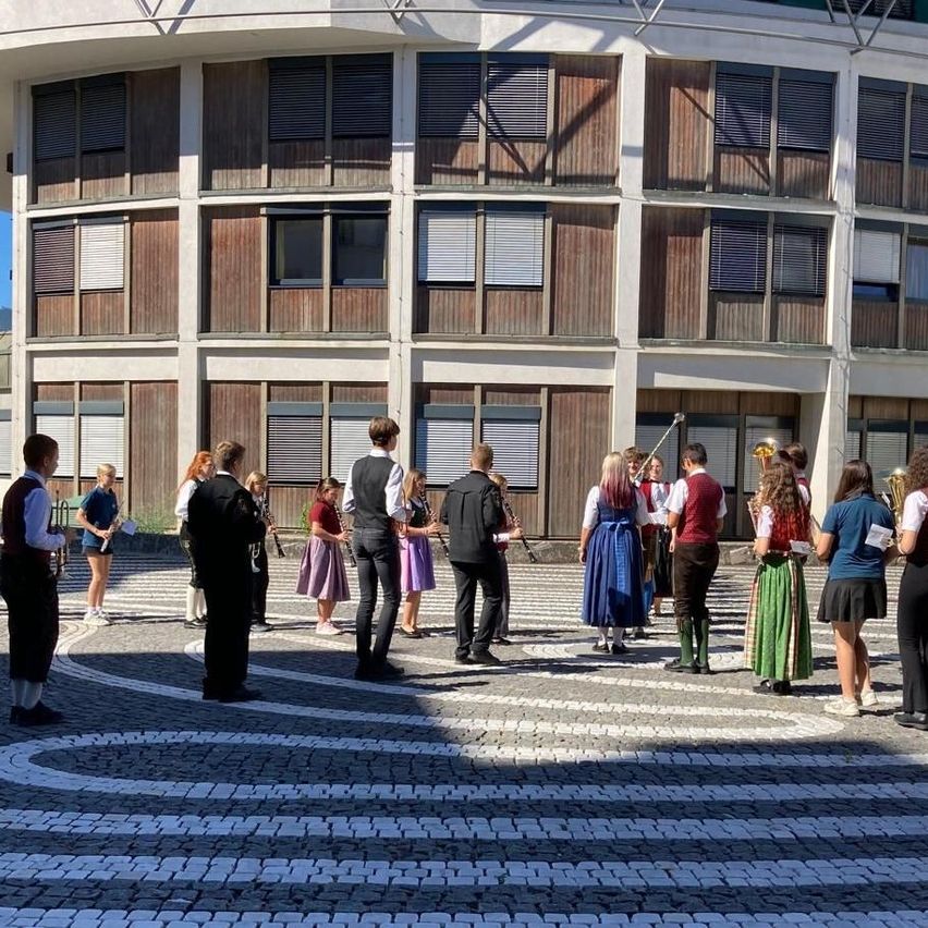 A group of people in traditional attire are gathered in front of a modern building, some holding musical instruments.