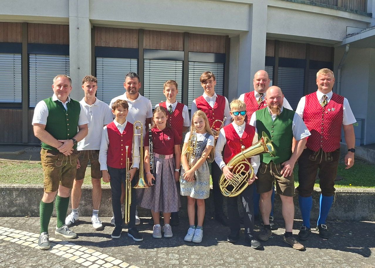 A group of musicians, both adults and children, dressed in traditional attire, are posing for a photo in front of a building. They hold various instruments like trumpets, trombones, and a tuba.