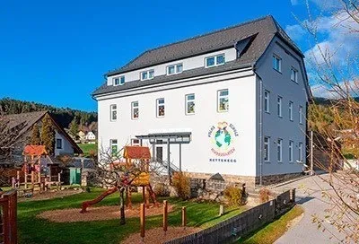 Ein weißes Gebäude mit mehreren Fenstern und einem grauen Dach verfügt über einen Spielplatz mit Rutsche und Klettergerüst. Das Gebäude trägt ein Schild mit 'SCHULEN' und 'KINDERGARTEN'. Im Hintergrund befinden sich Häuser und Bäume.