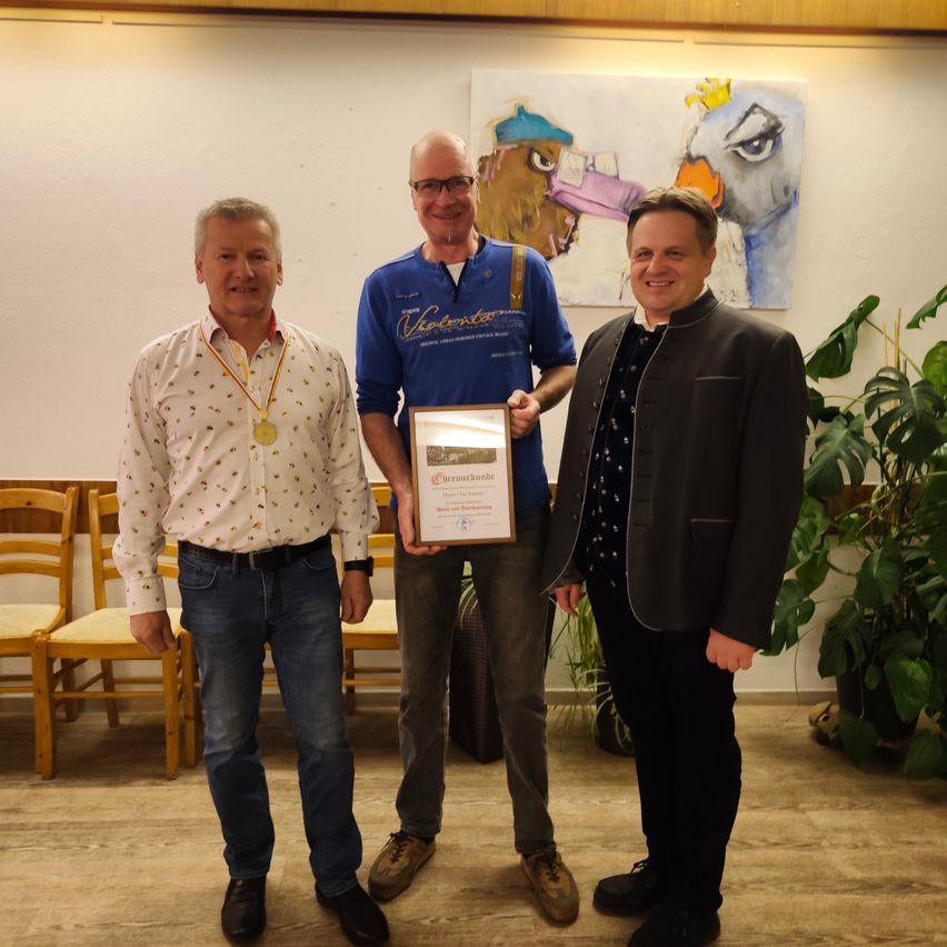 Drei Männer stehen lächelnd zusammen. Der Mann auf der linken Seite hat eine Medaille um den Hals. Der Mann in der Mitte hält ein gerahmtes Diplom. Der Mann auf der rechten Seite trägt einen dunklen Anzug. Sie befinden sich in einem Raum mit einem Bild an der Wand und Stühlen.