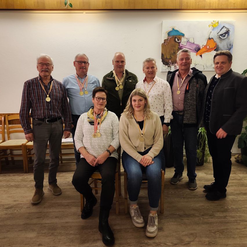 Eine Gruppe von acht Personen, darunter Männer und Frauen, mit Medaillen, posiert für ein Foto in einem Raum mit Holzböden und einer Wandmalerei.