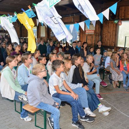 Eine Gruppe von Kindern und Erwachsenen sitzt auf Bänken in einem Raum. Einige tragen Brillen und Turnschuhe. Es hängen Kleidungsstücke und Fahnen. An der Wand befindet sich ein Banner mit dem Wort 'Miele' und ein Plakat.