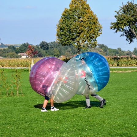 Zwei Personen auf einem Rasenfeld tragen aufblasbare Bälle, einer rosa und einer blau, mit Bäumen und einem Maisfeld im Hintergrund.