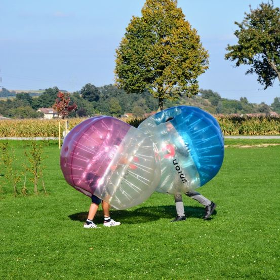 Zwei Personen auf einem Rasenfeld tragen aufblasbare Bälle, einer rosa und einer blau, mit Bäumen und einem Maisfeld im Hintergrund.