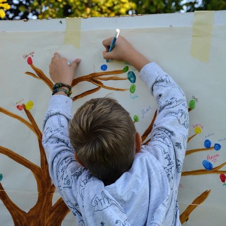 Ein junger Junge malt auf eine große Baumwand. Er hält einen blauen Stift und fügt den Ästen einen bunten Punkt hinzu. Die Wand ist weiß mit braunen Ästen und mehrfarbigen Punkten.