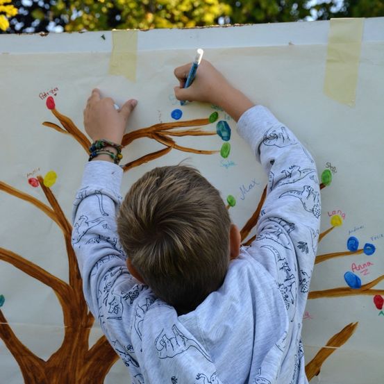 Ein junger Junge malt auf eine große Baumwand. Er hält einen blauen Stift und fügt den Ästen einen bunten Punkt hinzu. Die Wand ist weiß mit braunen Ästen und mehrfarbigen Punkten.