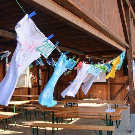Eine Holzstruktur mit Kleidern an einer Wäscheleine, darunter ein weißes T-Shirt mit dem Jahr 2012, ein blaues T-Shirt mit dem Jahr 2014 und weitere Shirts. Der Raum hat Holztische und -bänke.
