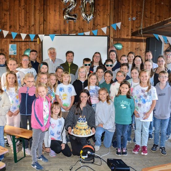 Eine Gruppe von Menschen, einschließlich Kindern, versammelt sich drinnen zu einer Geburtstagsfeier. Eine Frau hält einen Kuchen mit der Nummer 20, umgeben von lächelnden Gesichtern. Der Raum hat Holzwände und eine Tafel.