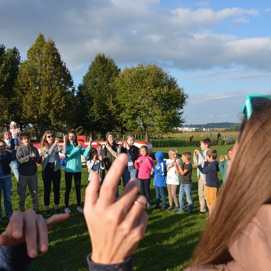 Eine Gruppe von Kindern und Erwachsenen versammelt sich auf einem Rasenfeld. Einige klatschen, während andere Kameras halten. Dahinter stehen mehrere Bäume, ein Gebäude und ein bewölkter Himmel.