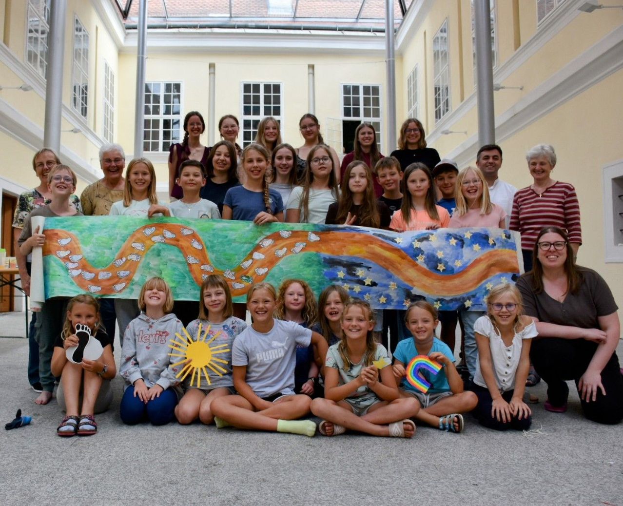 Eine Gruppe junger Mädchen und Jungen, zusammen mit ihren Lehrern, posiert für ein Foto in einem Flur. Sie halten ein langes Banner, das mit verschiedenen Elementen bemalt ist.