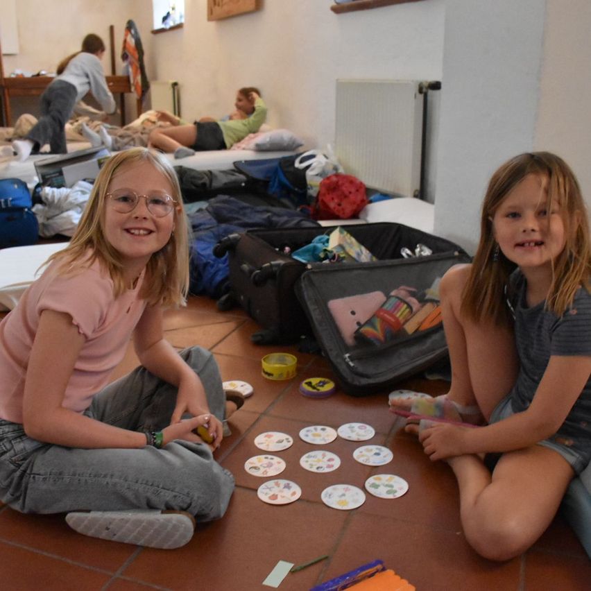 Zwei junge Mädchen sitzen auf dem Boden und spielen ein Spiel mit kleinen Karten. Sie lächeln. Hinter ihnen befinden sich Taschen, ein Bett und eine Frau, die auf einem Kissen liegt.