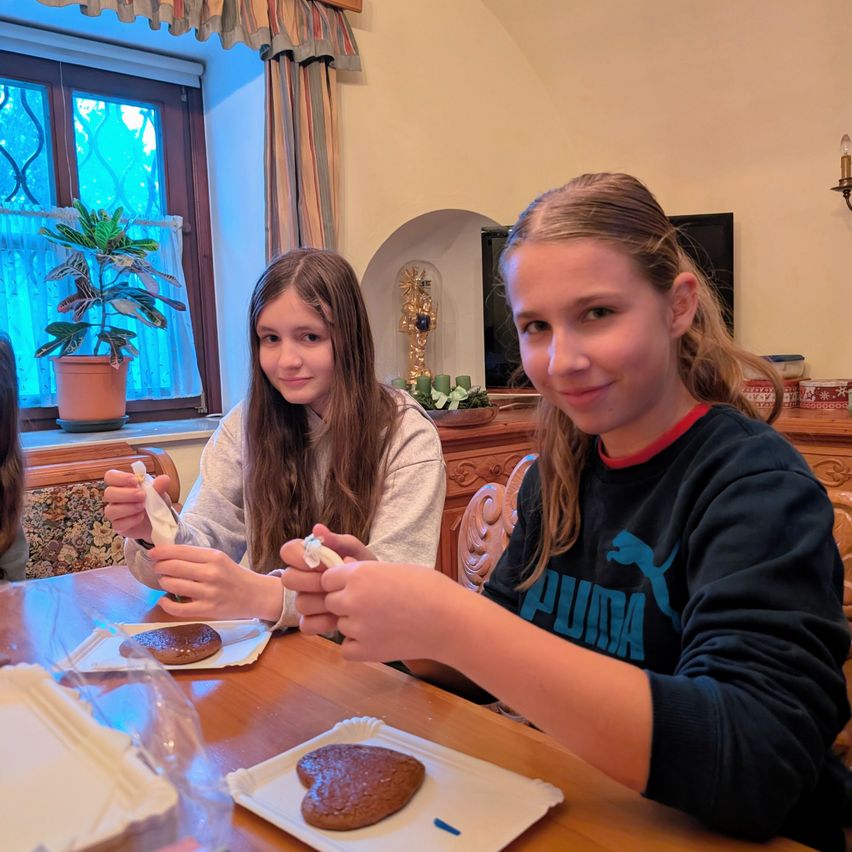 Zwei Mädchen sitzen lächelnd an einem Tisch in einem Zuhause. Sie halten ein kleines Objekt in den Händen. Eine Platte mit Keksen steht vor ihnen. Ein Topf mit einer Pflanze und ein Fenster sind hinter ihnen.