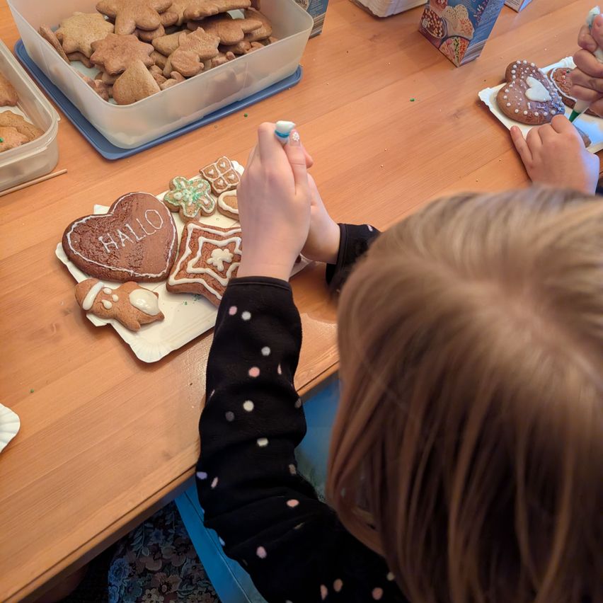 Ein Kind verziert Kekse auf einem Holztisch, mit verschiedenen Keksen und Zuckerguss auf dem Tisch. In der Nähe befindet sich eine Aufbewahrungsbox voller Kekse.