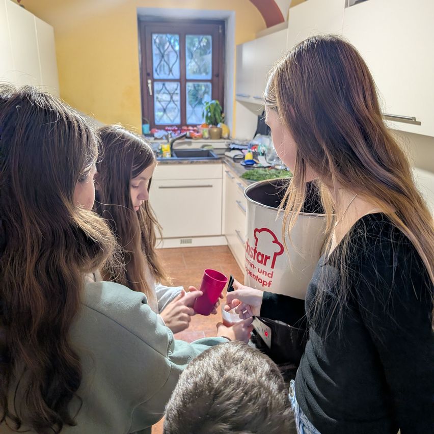 Vier Personen, drei Mädchen und ein Junge, sind in einer Küche. Sie stehen um einen großen Behälter herum und scheinen Essen zuzubereiten.