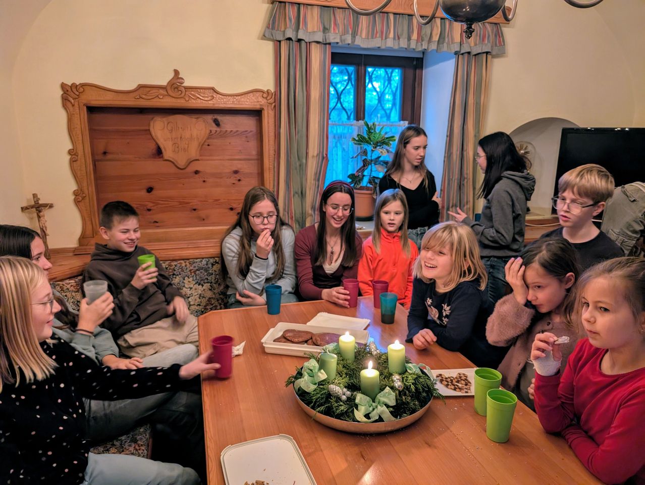 Eine Gruppe von Kindern und Erwachsenen sitzt um einen Tisch mit brennenden Kerzen und genießt Essen und Getränke. Der Raum hat Holzwände und Vorhänge.