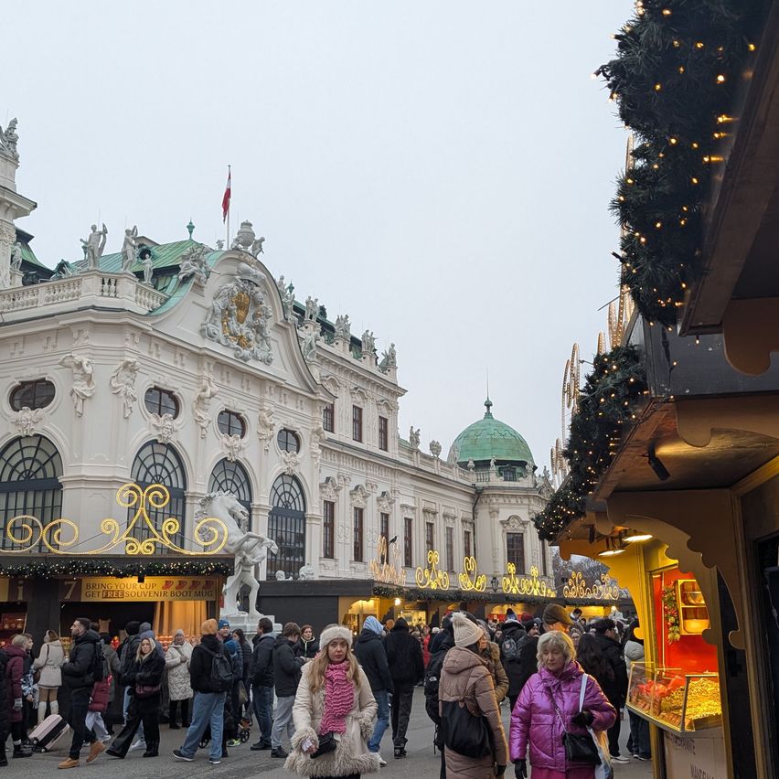 Eine Menschenmenge geht an einem geschmückten Gebäude mit einer Kuppel und einer Flagge vorbei. Vor ihm steht eine weiße Statue. Die Menschen sind in Winterkleidung gekleidet, und es gibt Weihnachtsdekorationen und Lichter.